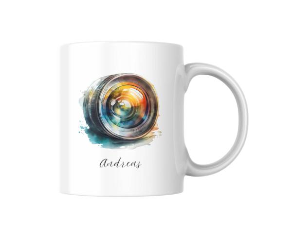 Eine weiße Keramiktasse mit einem bunten, aquarellierten Motiv eines Kameraobjektivs. Das Objektiv zeigt lebendige Farben und ist umgeben von Farbspritzern. Unten auf der Tasse steht der Name „Andreas“ in einer eleganten Schrift. Diese Tasse ist ein ideal